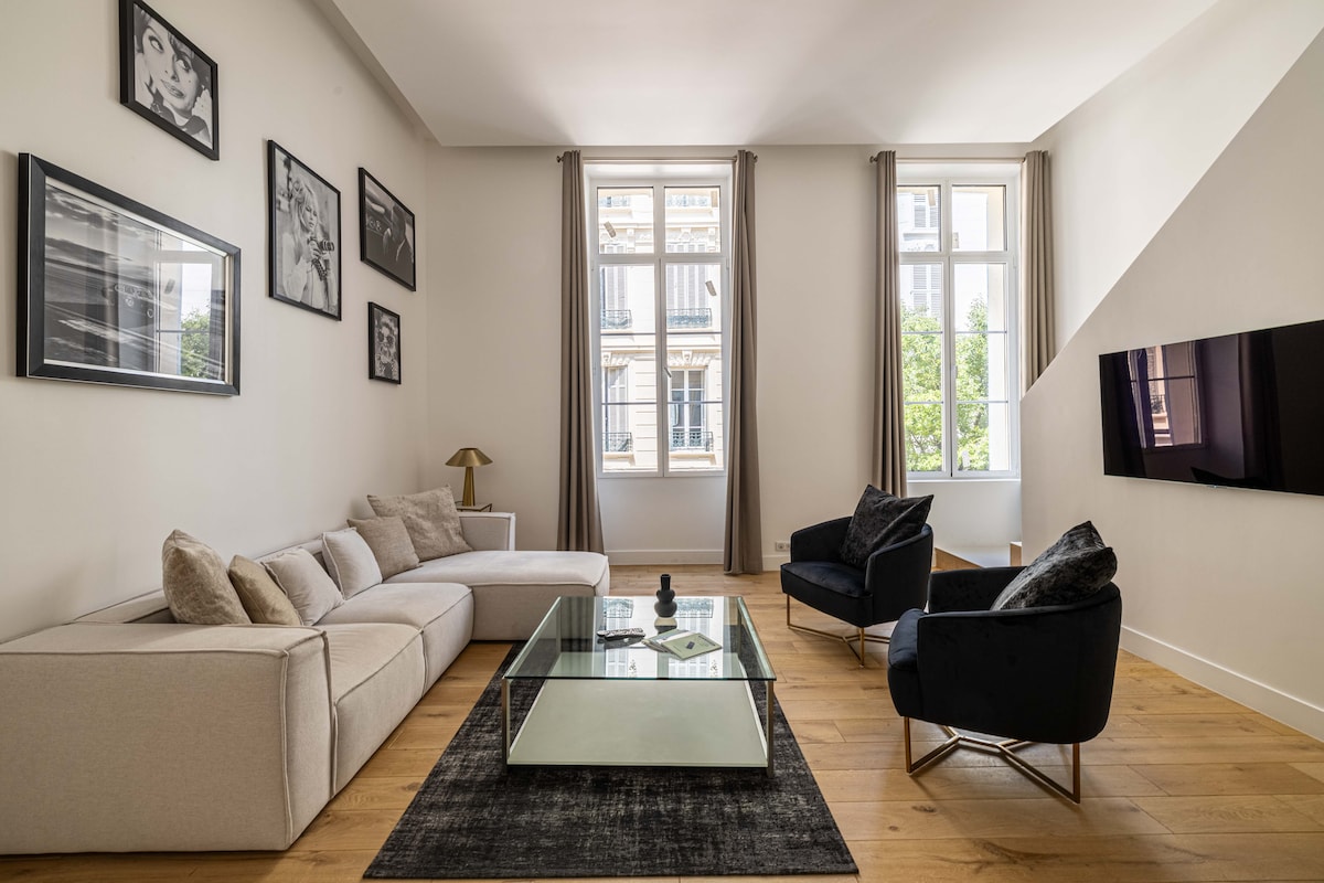 A spacious living area is furnished with a modern sectional sofa and two accent chairs. A glass coffee table sits in the center, and art pieces are displayed on the walls. Natural light brightens the room through large windows, creating a welcoming environment.