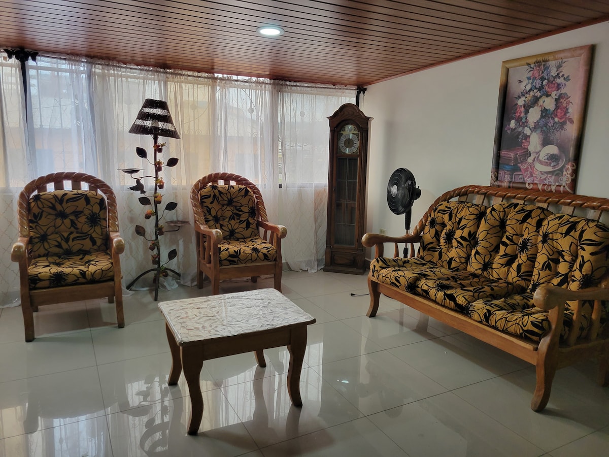 A bright, spacious living area features comfortable seating with floral upholstery. Two wooden chairs and a three-seater sofa surround a small coffee table. A vintage clock and a decorative lamp add character, while large windows allow natural light to fill the space.