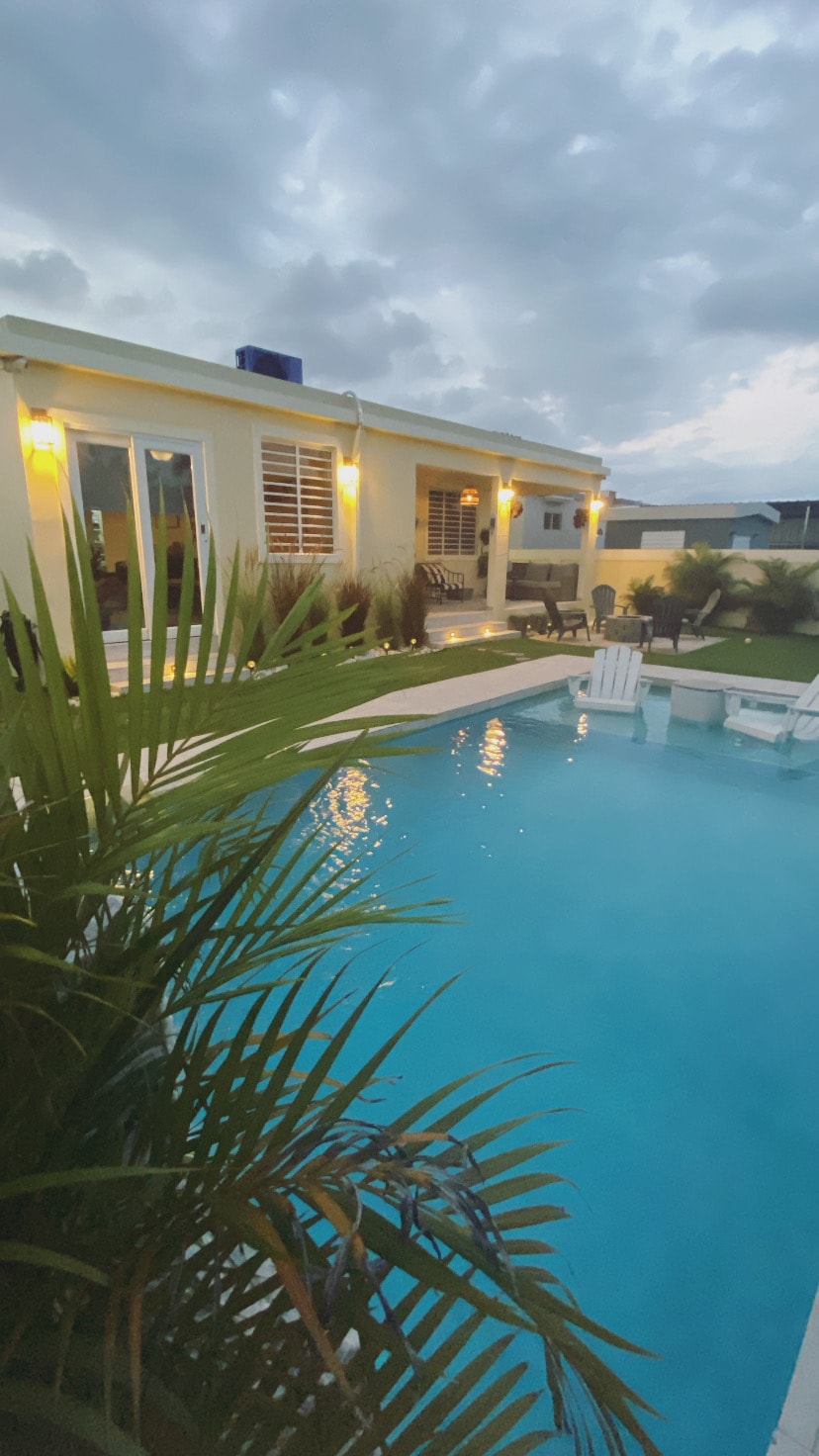 A serene outdoor pool area is framed by lush greenery, reflecting soft evening light. The pool is accompanied by a seating area featuring chairs and a table. The exterior of the house showcases large windows and subtle lighting, contributing to a relaxing ambiance.