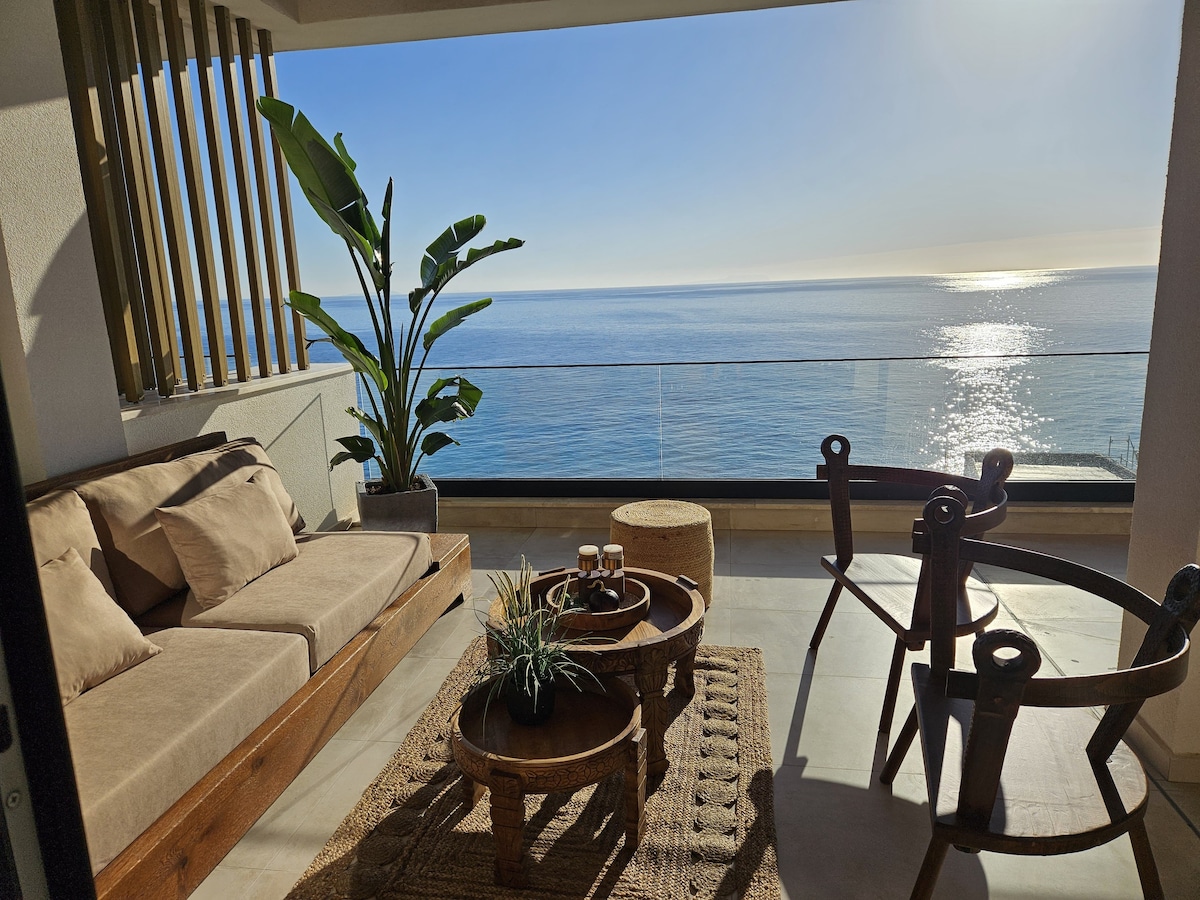 A spacious terrace is shown with modern seating arrangements facing the Ionian Sea. Natural light pours in through a large glass door, highlighting a comfortable sofa and a wooden coffee table adorned with decorative items. Potted plants add a touch of greenery to the serene outdoor space.