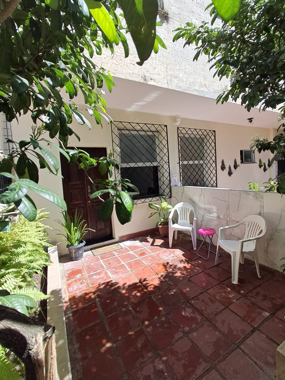 An outdoor area is surrounded by lush greenery, featuring a tiled floor and two white plastic chairs. A small pink chair adds a pop of color. The entrance is framed by a decorative fence, with sunlight filtering through the leaves, creating a peaceful ambiance.