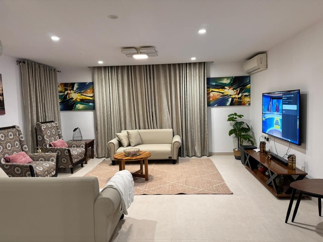 The living room features a comfortable sofa and two patterned armchairs arranged around a circular coffee table. Two colorful artworks adorn the walls, complemented by soft drapes that frame the windows. A flat-screen television is mounted on a wall unit, and a potted plant adds a touch of greenery.