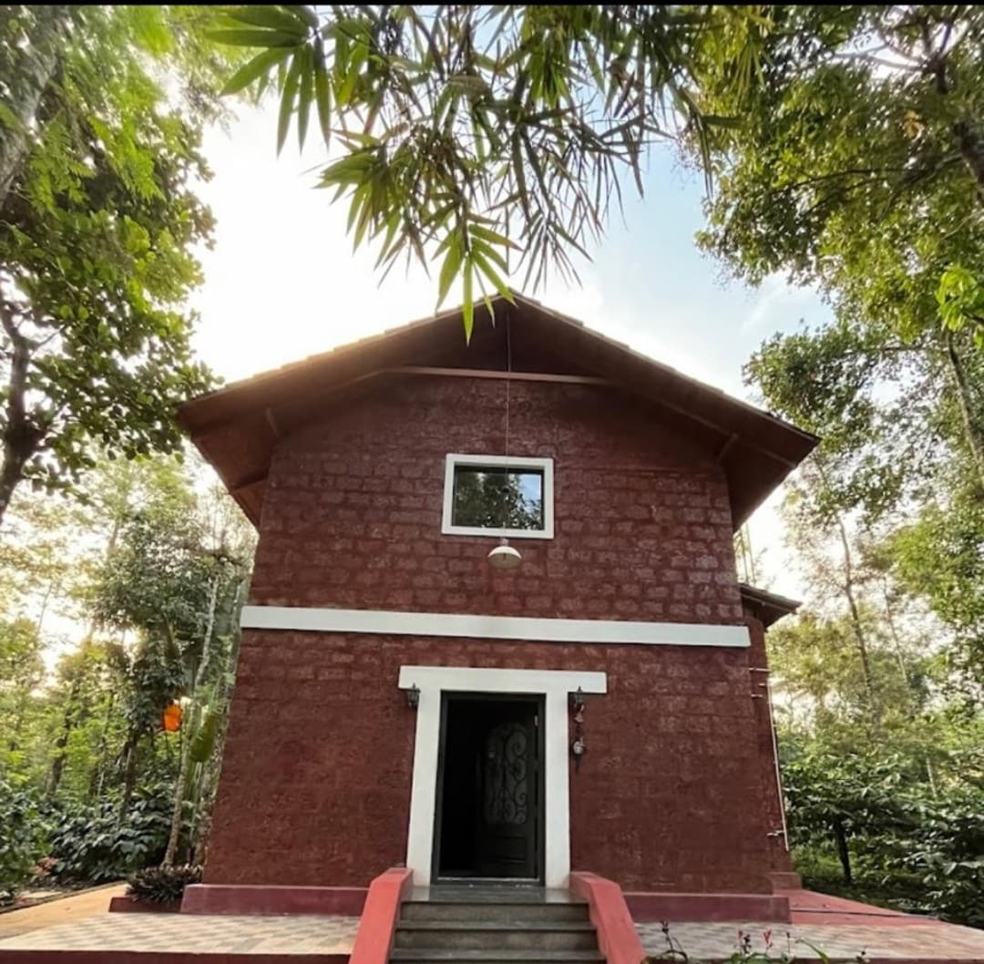 A two-story house is situated among lush greenery, featuring a textured red exterior. Large windows are positioned prominently, allowing natural light to enter. A welcoming front entrance is framed by steps leading up to the door, surrounded by vibrant foliage.