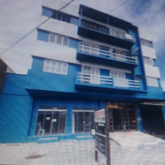 A multi-story building features a vibrant blue exterior with multiple balconies. Large windows provide ample natural light to the ground floor, where signage indicates the entrance. The surrounding area includes a path leading up to the building.