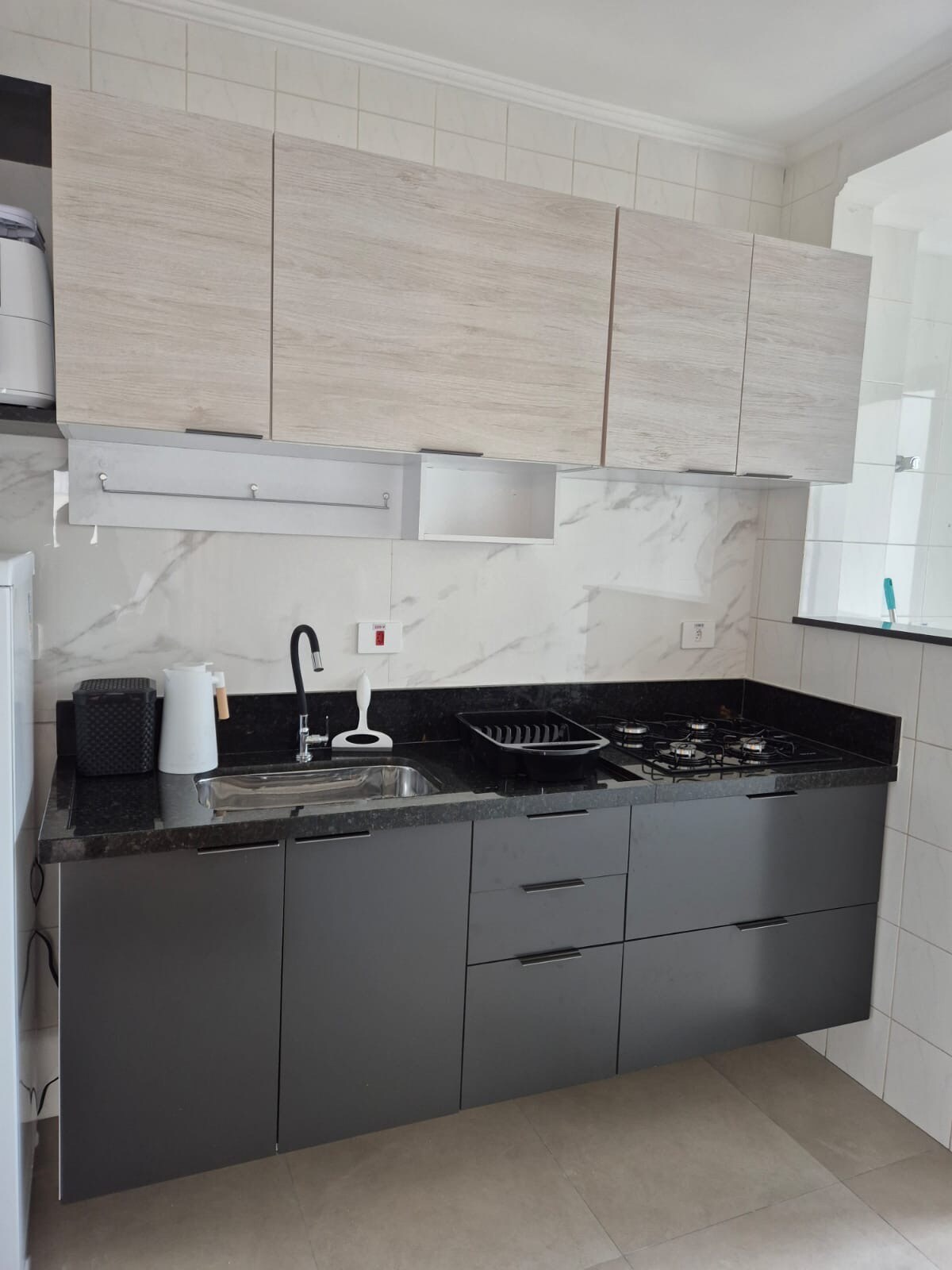 A modern kitchen setup is presented, featuring sleek cabinetry in light and dark tones. Essential appliances like a refrigerator and a stove are visible. A black countertop holds a sink and kitchen utensils, with ample storage options above and below.