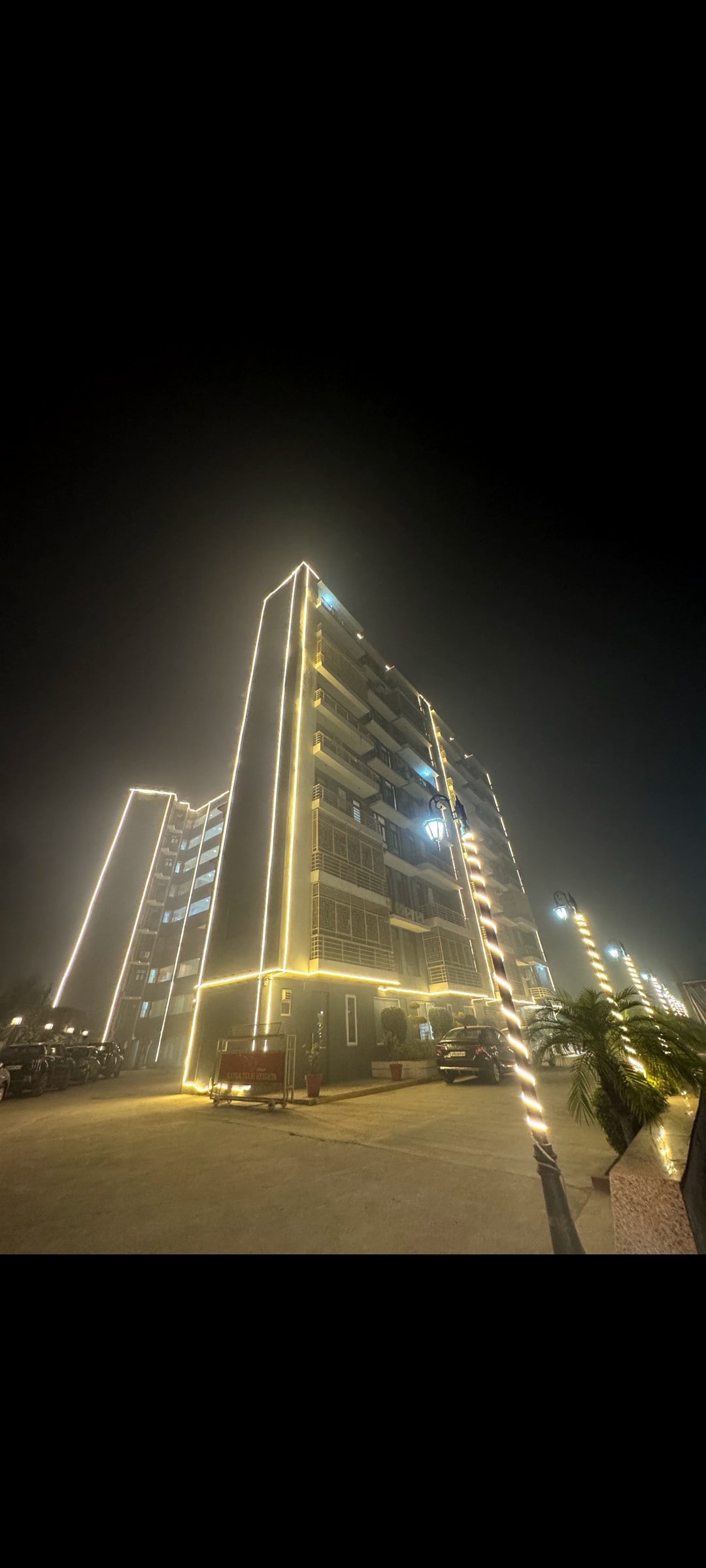 The exterior of a modern building is illuminated with soft lights, creating a warm ambiance at night. The structure features multiple balconies and large windows, while surrounding parking areas and palm trees are visible, enhancing the welcoming atmosphere.