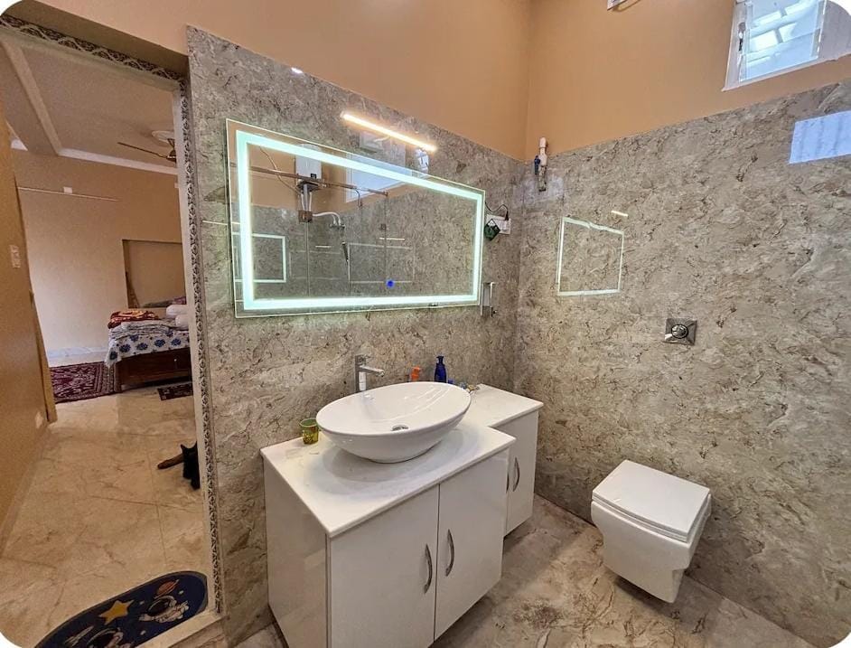 A modern bathroom features a circular sink atop a white vanity, complemented by a large illuminated mirror. Smooth marble walls and floors contribute to a clean, spacious feel. A toilet and small shelf are positioned to the right, with natural light filtering in from a window.