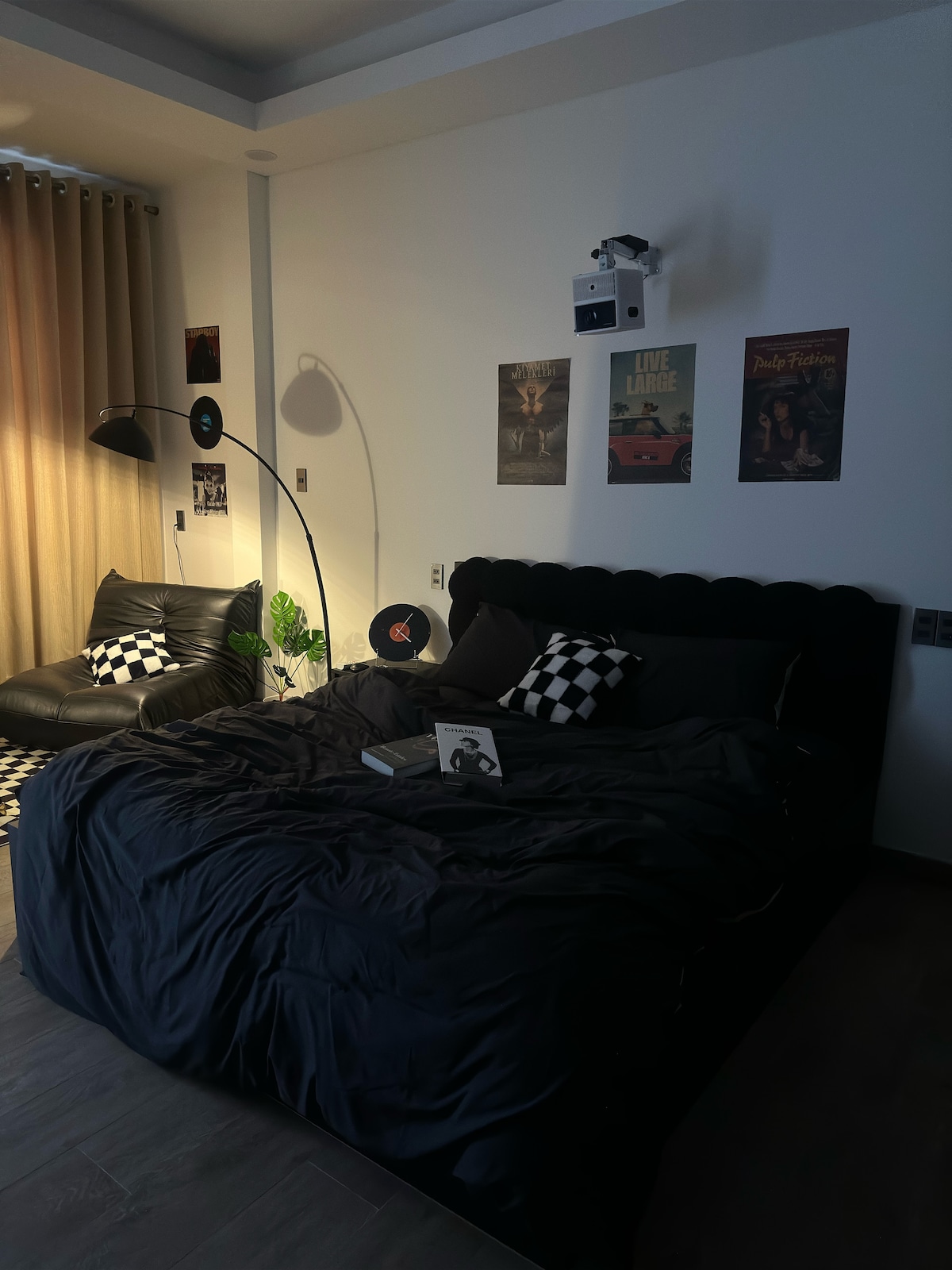 A peaceful sleeping area features a spacious bed with a textured black duvet and patterned throw pillows. An arched floor lamp provides soft lighting, while framed artwork adorns the walls. A cozy chair and a small green plant add a touch of comfort to the space.