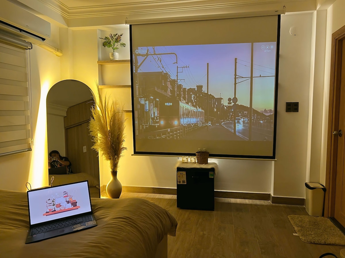 A modern room features a spacious bed with soft linens, a laptop displaying a colorful image, and a large projection screen showcasing a sunset scene. Ambient lighting adds warmth, while decorative elements include potted plants and a minimalist coffee table.