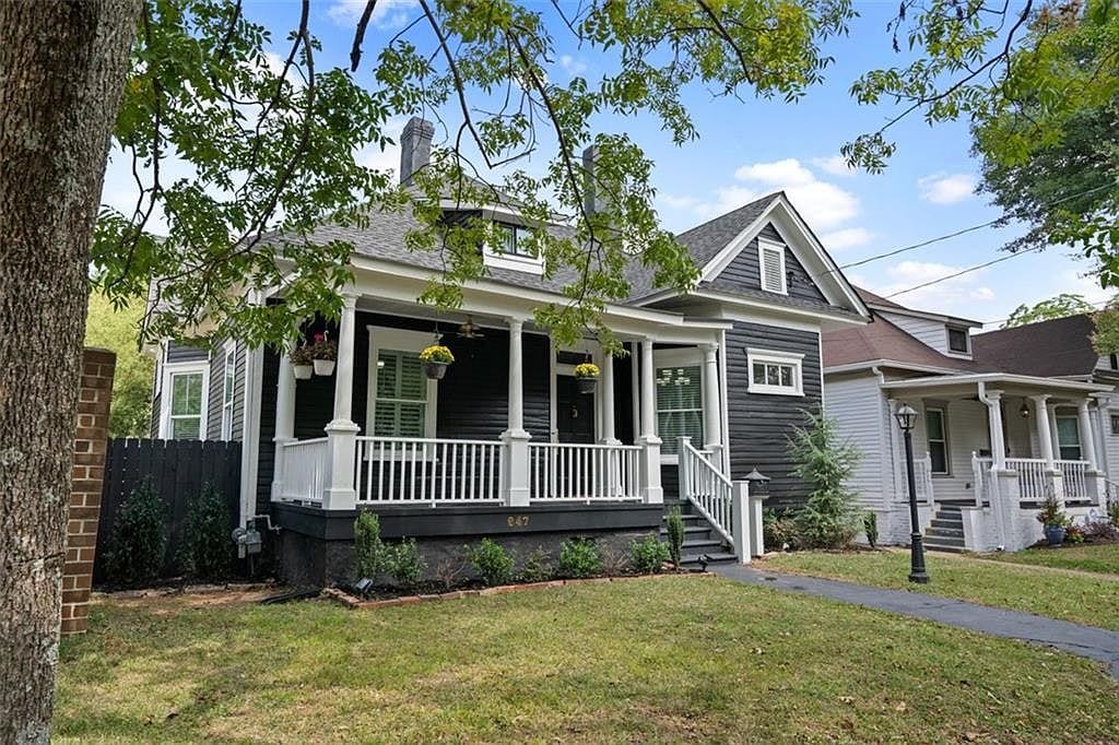 A charming exterior of a historic home is featured, with a welcoming porch and white railing. Lush greenery surrounds the property, including hanging flower pots. A pathway leads up to the entrance, framed by soft grass and shade from nearby trees.