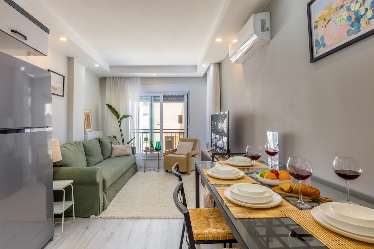 A modern living area is featured, showcasing a spacious sofa and an armchair, with a glass-top dining table set for four. A flat-screen TV is positioned nearby, while a large window invites natural light. A potted plant adds a touch of greenery to the space.