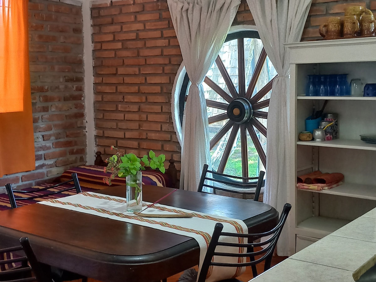 A dining area is showcased with a wooden table and several black chairs, accompanied by a decorative table runner. A large circular window resembling a wagon wheel allows natural light to filter in, with greenery visible outside and rustic brick walls adding texture to the space.