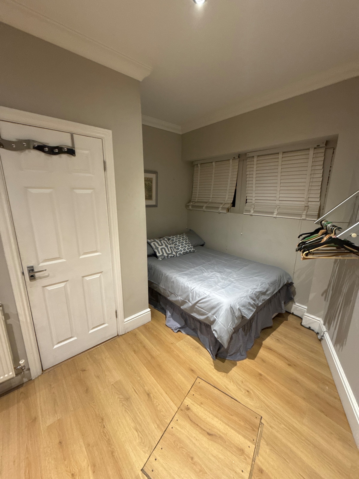 A compact bedroom features a neatly made bed with a light grey coverlet and decorative pillows. Natural light enters through two fitted window blinds. A small closet with hangers is situated beside the door, and wooden flooring enhances the room's warmth.