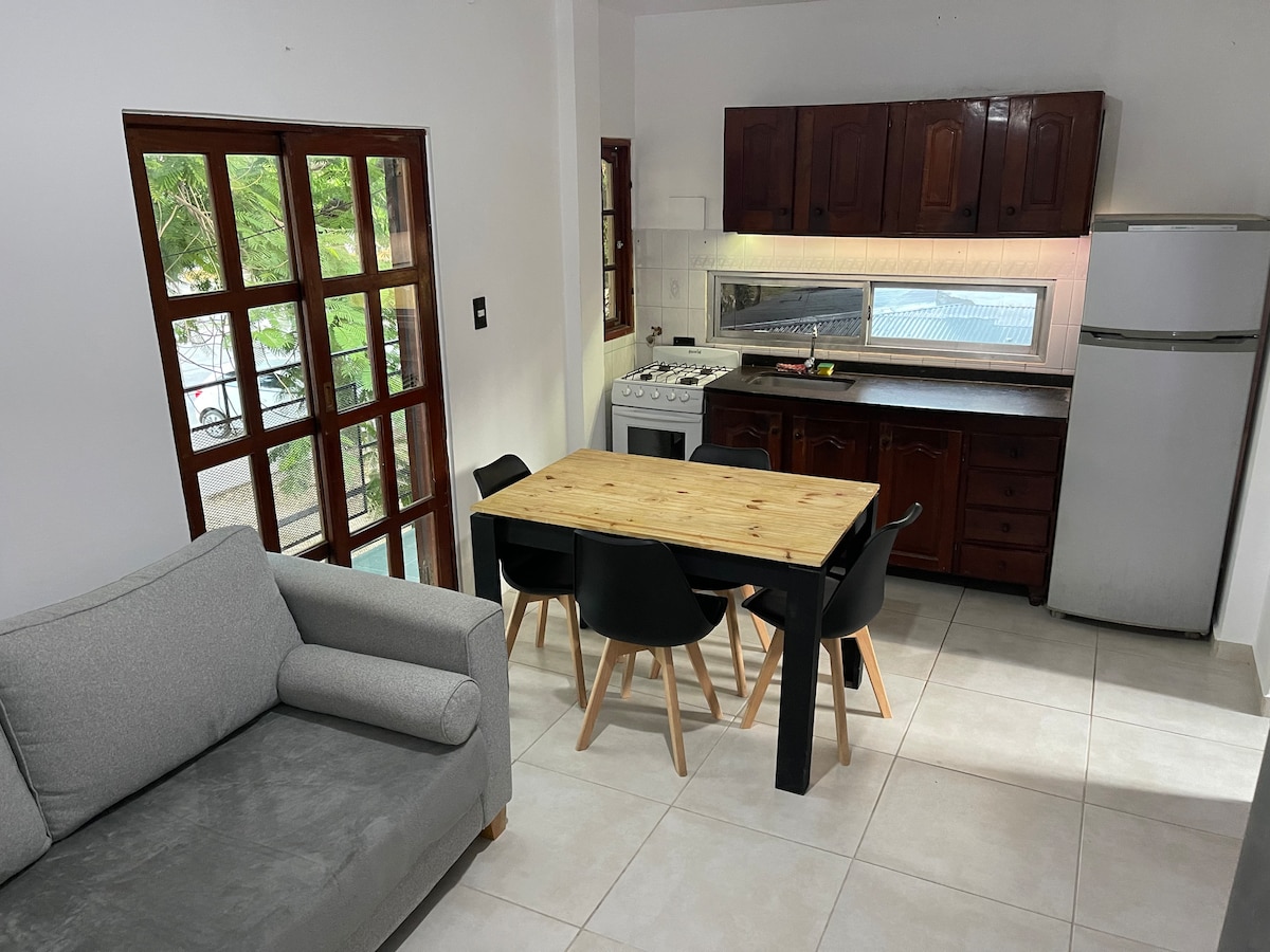 An open kitchen area is visible, featuring a wooden dining table surrounded by black chairs. The kitchen is equipped with a stove, refrigerator, and ample counter space. Natural light enters through large windows, illuminating the adjacent cozy seating area.