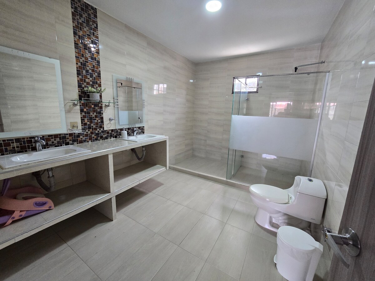 A spacious bathroom is equipped with two sinks positioned on a broad countertop. A glass-enclosed shower area is visible on one side, while a white toilet is placed on the opposite side. Neutral-colored tiles adorn the walls and floor, creating a clean and modern look.