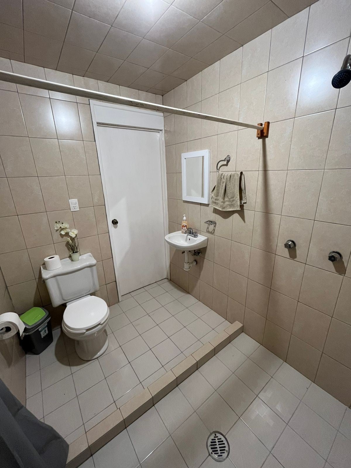 A neatly arranged bathroom is visible, featuring beige tiled walls and floors. A white toilet and a small sink are positioned against the wall. A towel hangs from a hook, and a shower area with a curtain is seen in the corner. A mirror reflects the light.