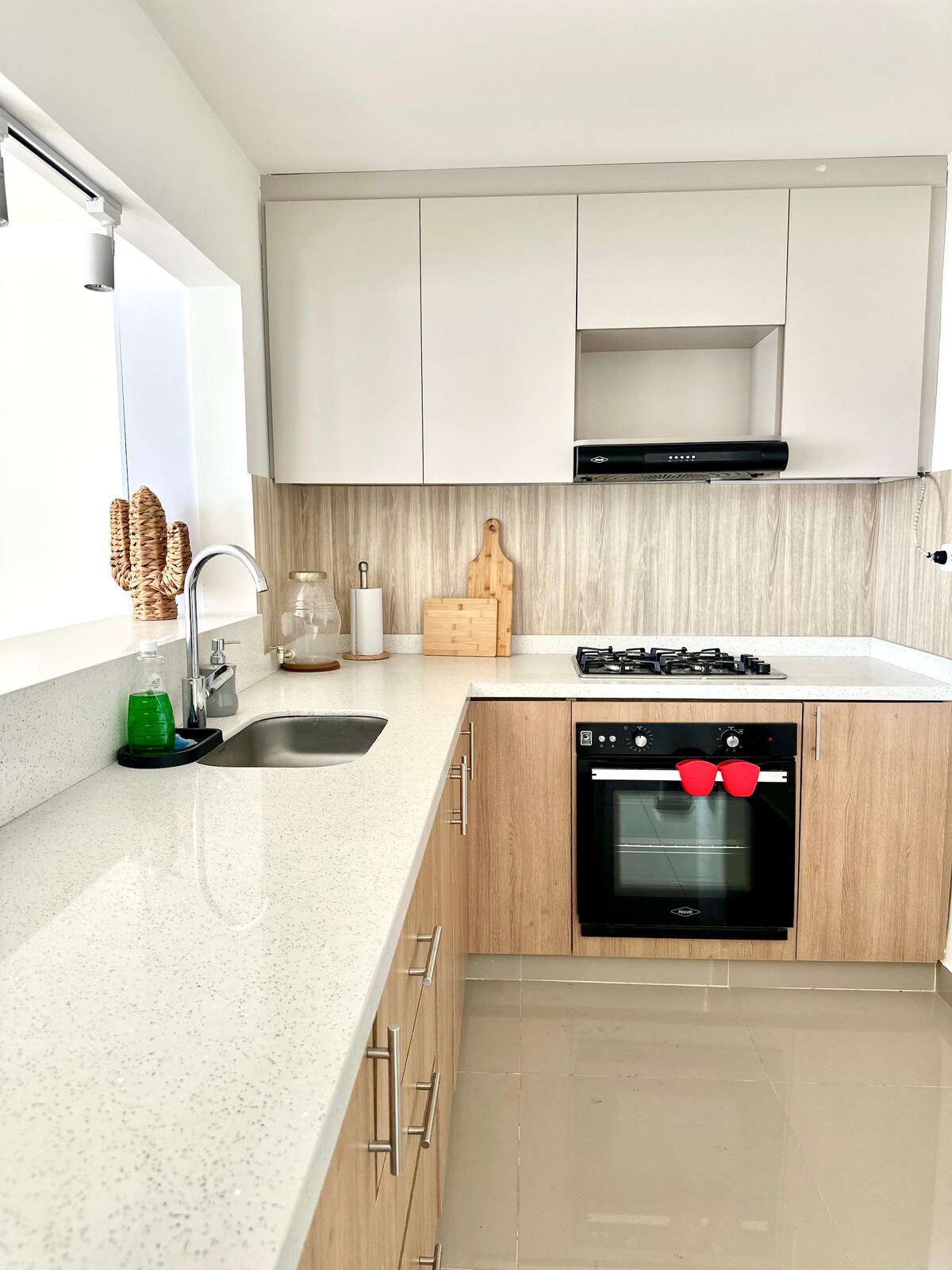 A modern kitchen features sleek cabinets in light tones, complemented by a spacious countertop. A gas stove is positioned next to a sink, with a decorative cutting board displayed on the wall. Natural light filters through a window, illuminating the thoughtfully designed space.