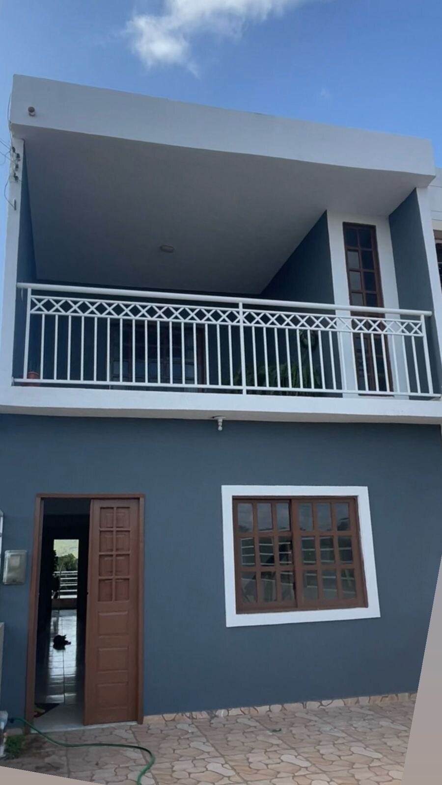 A two-story house is portrayed, featuring a light blue exterior with white accents. A balcony is visible on the upper floor, enclosed by a decorative railing. Large windows are present on both levels, promoting natural light. The entrance door is textured wood, inviting guests inside.