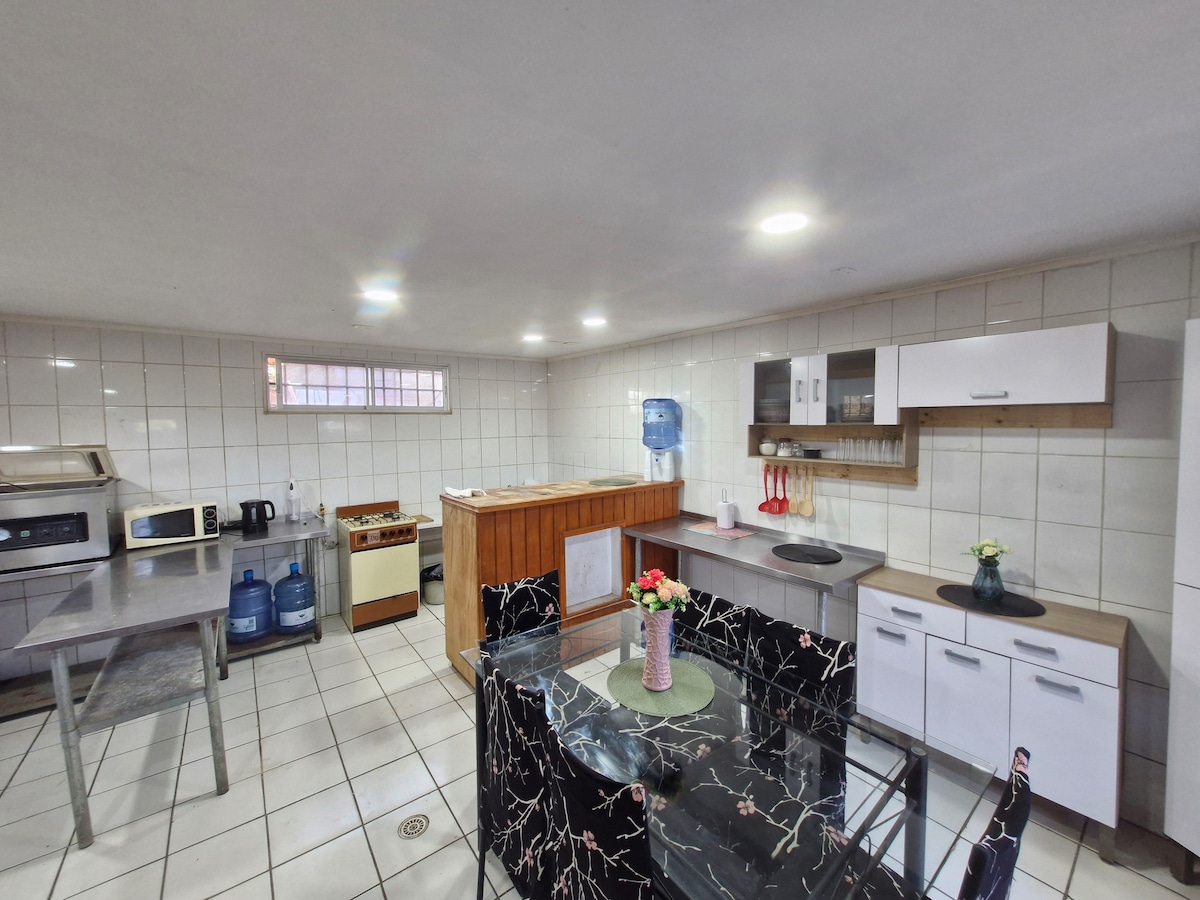 A spacious kitchen area features a variety of appliances, including a microwave and a cooking stove. Sleek countertops provide ample workspace. A dining table with floral centerpiece is positioned beside the cabinetry. Natural light floods through a window, enhancing the kitchen's functionality.
