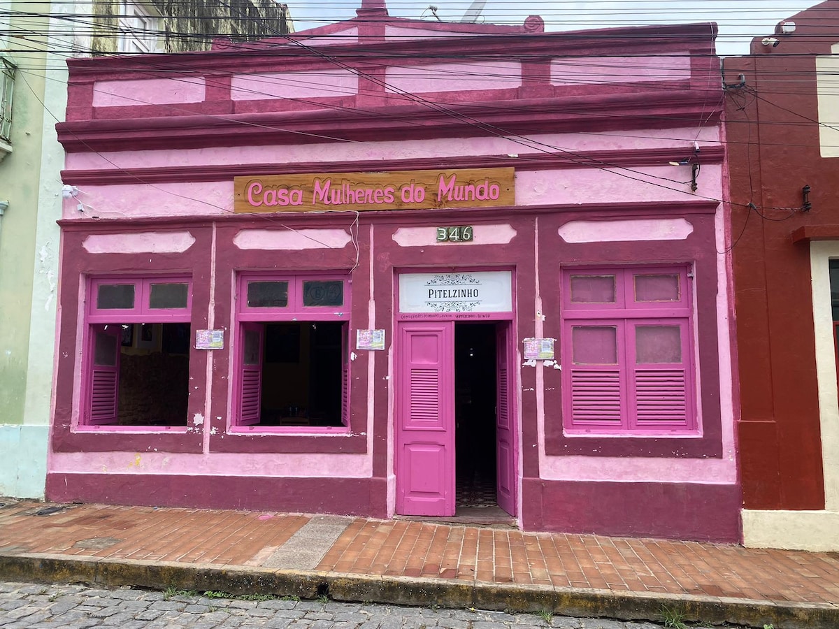 A vibrant pink exterior is showcased, featuring large windows framed in lighter shades of pink. The establishment is identified by the sign 'Casa Mulheres do Mundo,' above the entrance, which includes a door painted in matching pink tones. The sidewalk is lined with warm terracotta tiles.