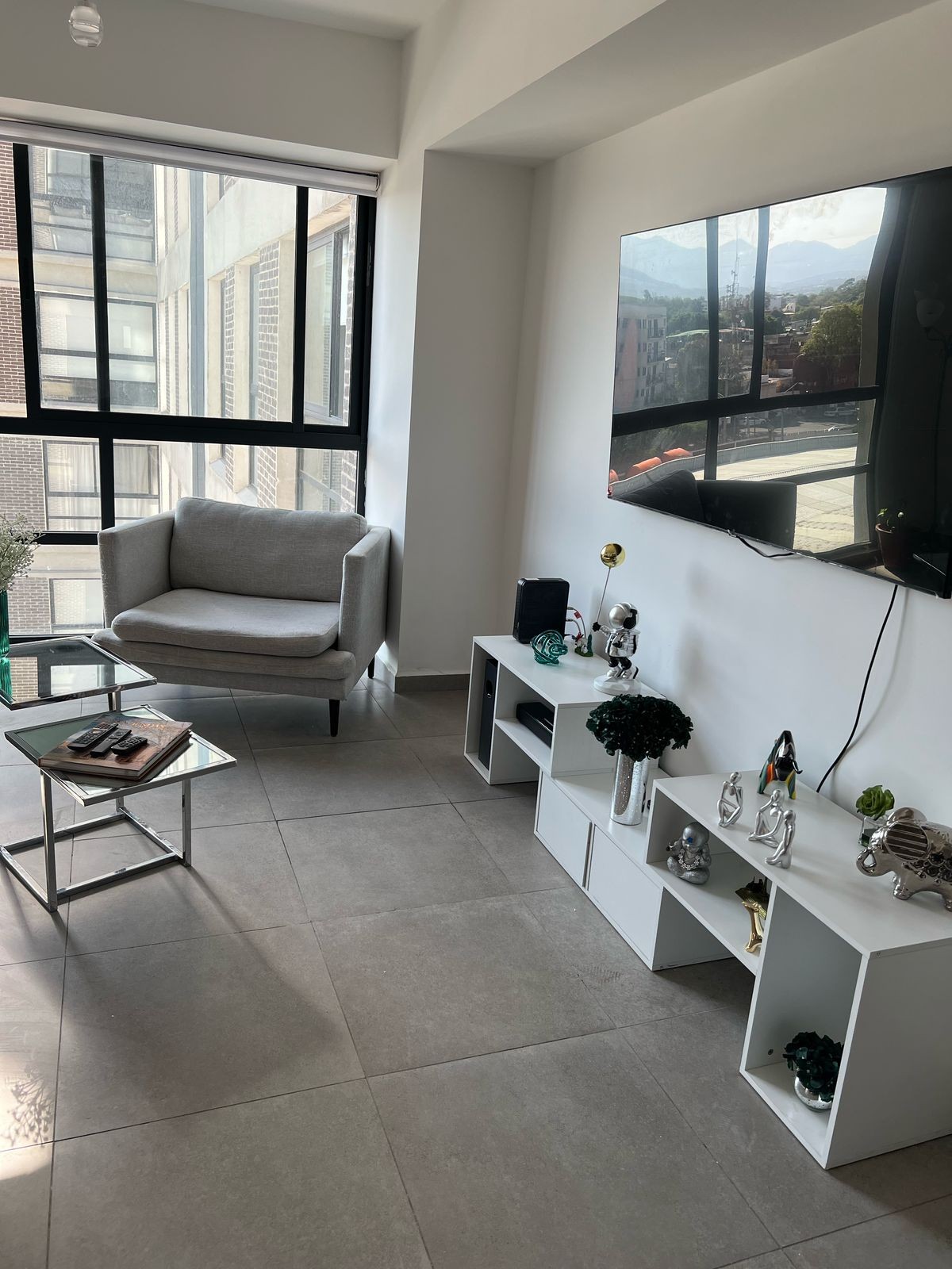 A well-lit living area is shown, featuring a gray armchair positioned beside large windows. A sleek glass coffee table is displayed, and a low white shelf holds various decorative items. A wall-mounted TV dominates one side, enhancing the room's modern design.