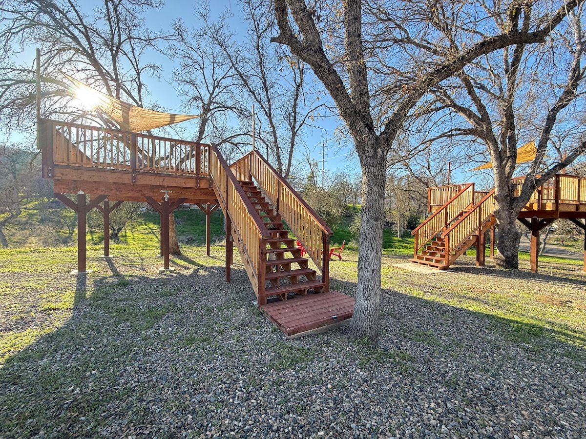 Wooden tree decks are positioned among bare oak trees, featuring sturdy staircases leading to elevated platforms. Dappled sunlight illuminates the scene, casting soft shadows on the gravel ground. The area is surrounded by lush greenery, enhancing the natural ambiance.