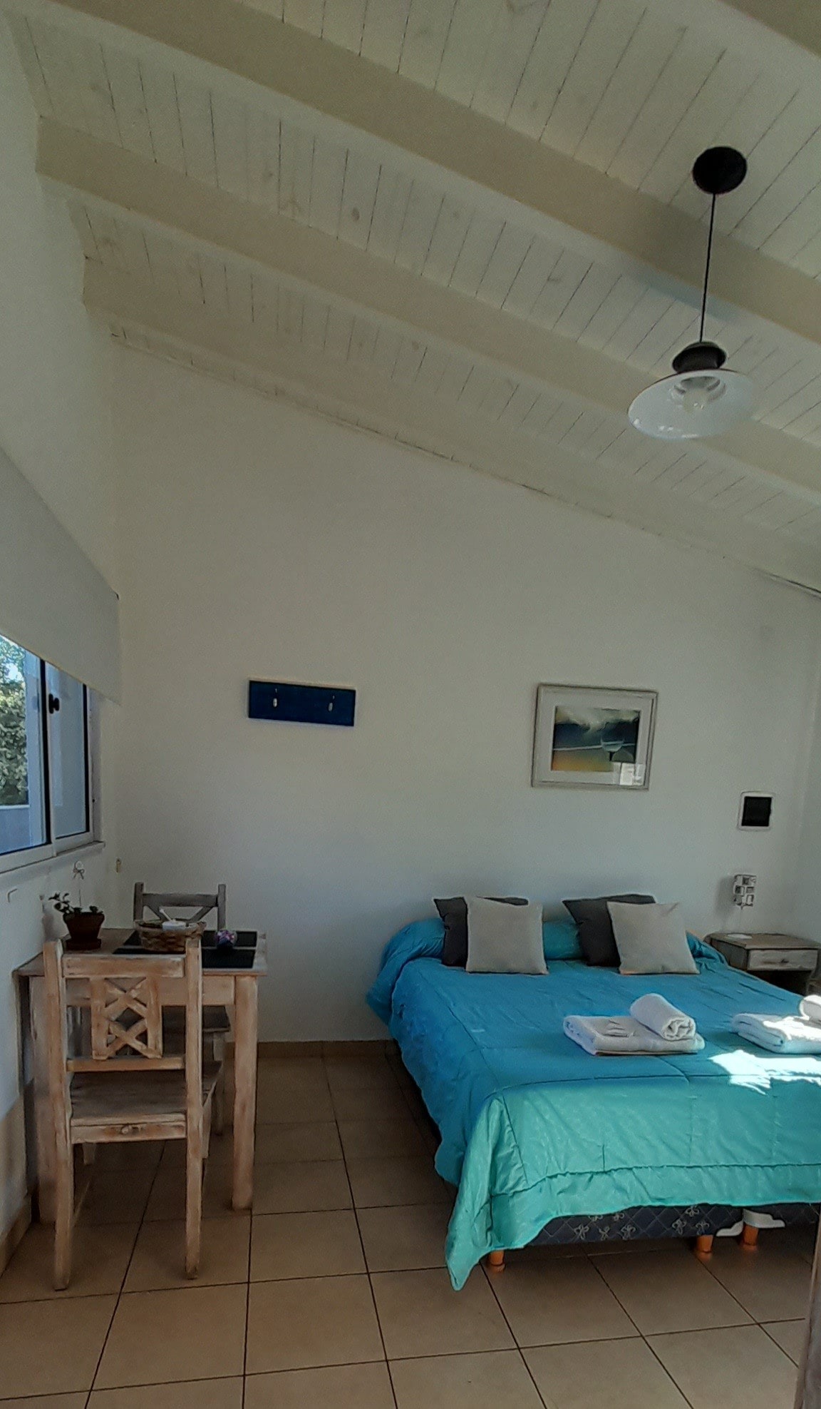 A well-appointed room features a spacious bed covered with a turquoise blanket, complemented by soft pillows. A simple wooden table and chair set is positioned nearby, while a framed artwork hangs on the wall. Natural light enters through a window, enhancing the bright atmosphere.
