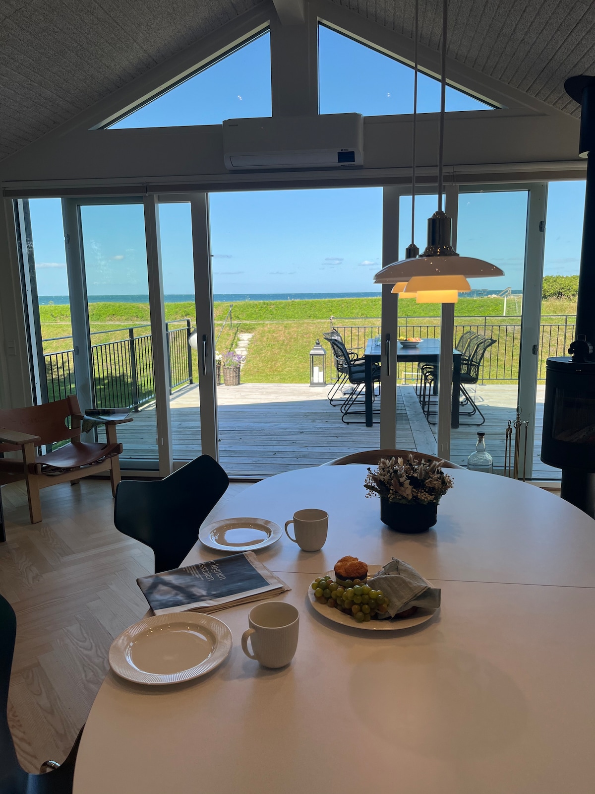 The dining area features a circular white table set with plates, cups, and a small basket of fruit. Large glass doors offer a view of the outdoor terrace and sea, allowing natural light to fill the space.