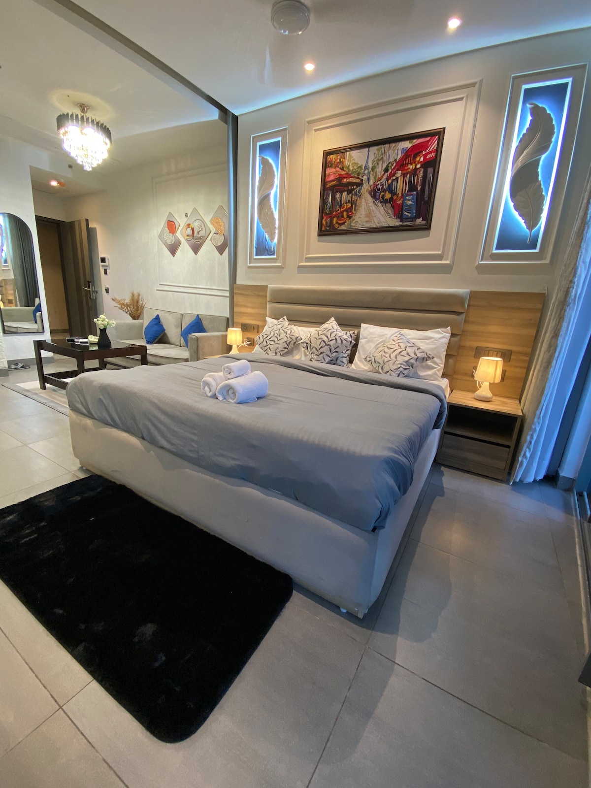 A spacious sleeping area features a comfortable bed with soft linens and decorative pillows. A modern artwork hangs above the bed. Two blue cushions are placed on a small seating area with a low table, and a plush black rug adds warmth to the floor.