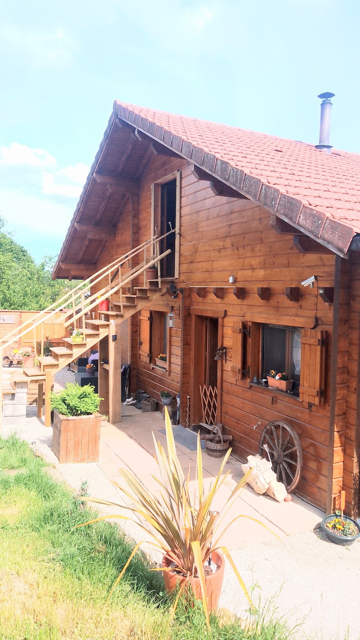 The exterior of a charming wooden chalet is presented, featuring a sloped roof and a set of stairs leading to a second entrance. Planters with plants are visible, and rustic elements such as a decorative wagon wheel enhance the welcoming atmosphere of the space.