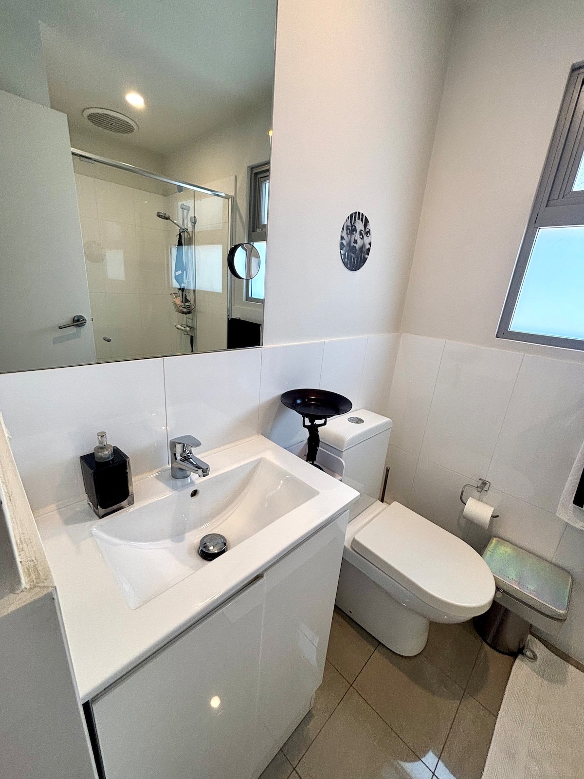 A compact bathroom is equipped with a modern sink and a sleek toilet. A large mirror extends above the sink, reflecting the light from nearby windows. The tiled floor adds a polished look, while a shower area is visible through a glass door.