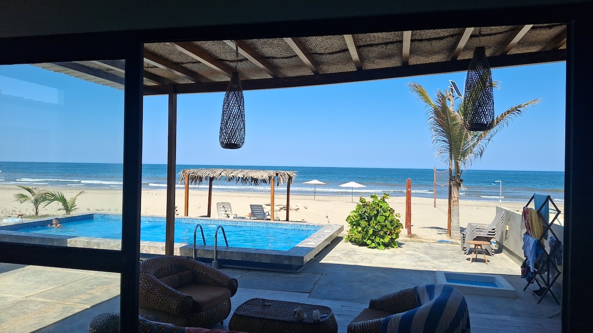 The image captures a beachfront view from inside the property, featuring a calming infinity pool on the left. Palm trees and sun loungers dot the sandy beach, while a cabana with a thatched roof stands in the foreground. A clear blue sky complements the serene ocean.