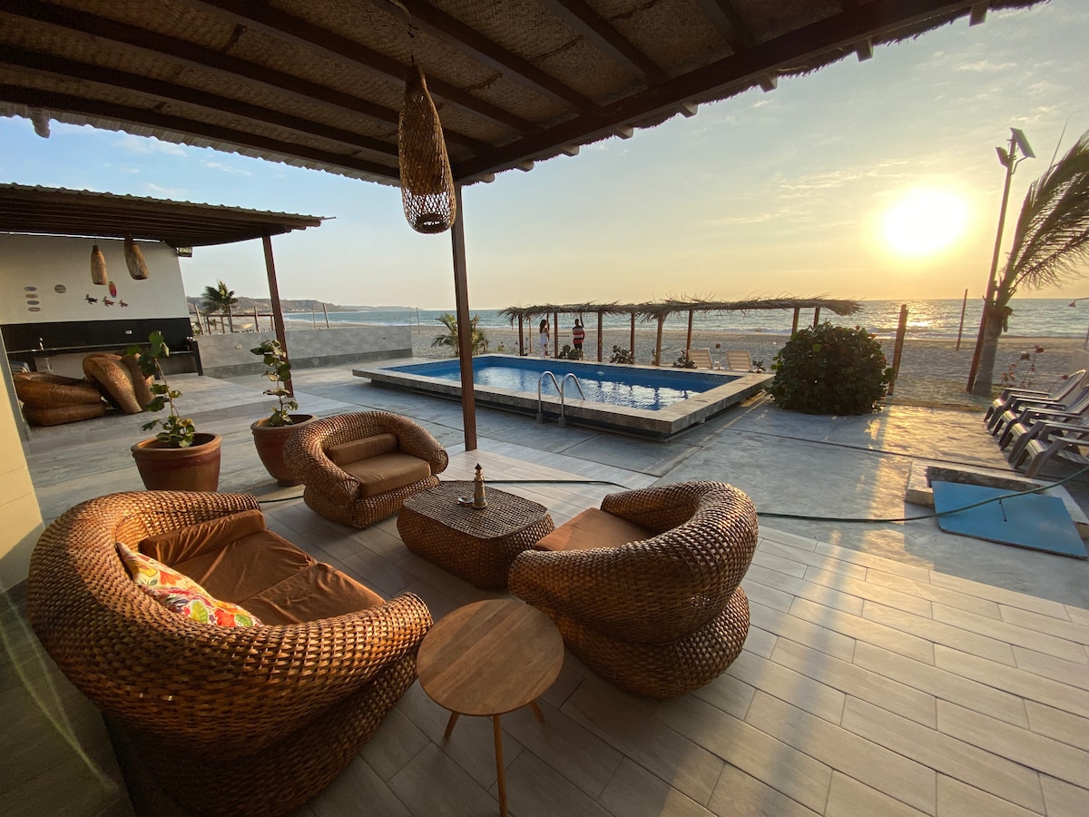 An outdoor seating area features woven lounge chairs and a wooden table, positioned under a thatched roof. A pool is located nearby, with the beach and ocean visible in the background. The sunset casts a warm glow over the entire scene.