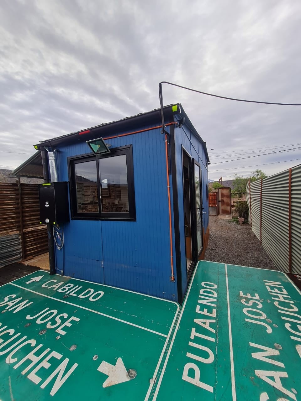 A compact blue structure is set against a cloudy sky, featuring large windows that allow natural light. Green directional signs in the foreground indicate nearby locations, while gravel paths surround the building, emphasizing its unique placement in the landscape.