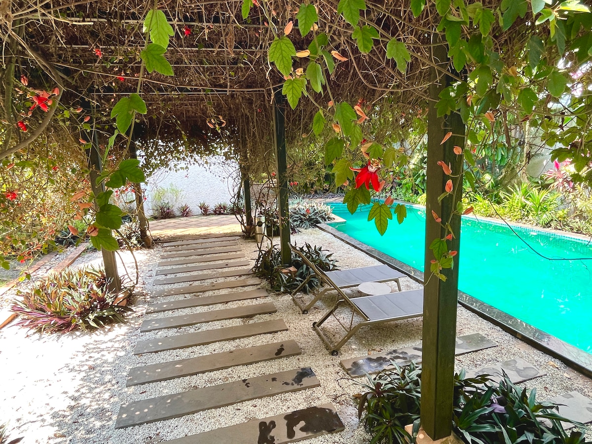 A pool area is framed by lush greenery and vibrant flowers, with a wooden structure overhead providing natural shade. Stone pathways lead towards a turquoise pool, bordered by neatly arranged plants, creating a serene retreat space.