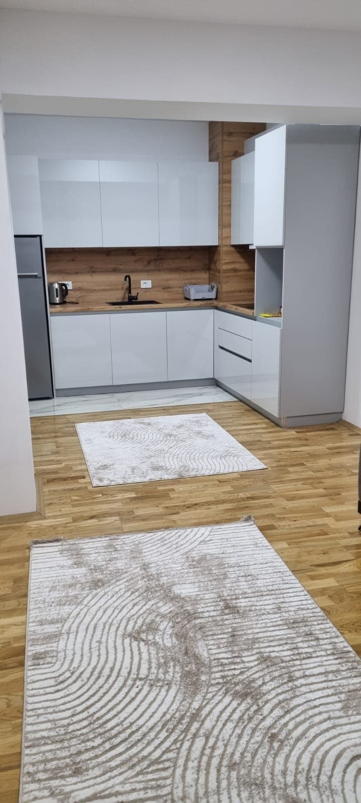 A modern kitchen features sleek white cabinetry and a wooden accent wall. Essential appliances, including a refrigerator and coffee maker, are visible. Two area rugs add warmth to the space, which is well-lit with natural light streaming in.