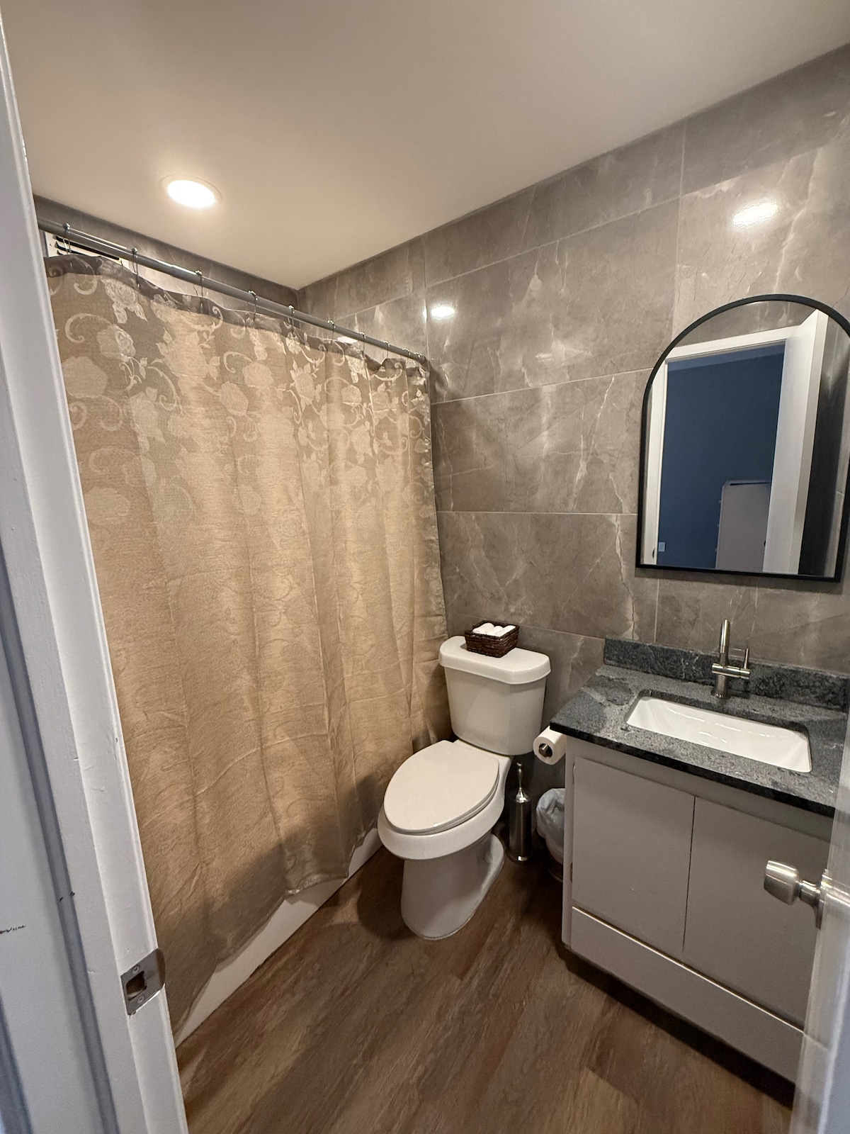 A modern bathroom is presented with grey tiled walls and a light-colored vanity. A shower area is separated by a textured curtain, and a compact toilet is positioned beside the vanity. Soft lighting enhances the sleek design of the space.