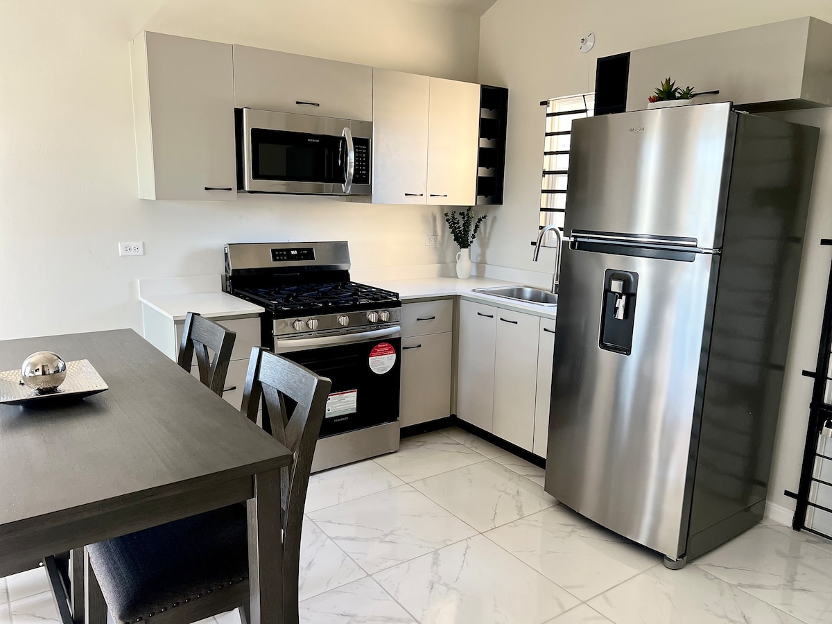 A modern kitchen is shown, equipped with stainless steel appliances including a gas stove, microwave, and refrigerator. The cabinetry features a sleek design, complemented by a dining table with four chairs. Bright white tiles adorn the floor, enhancing the spacious feel of the area.