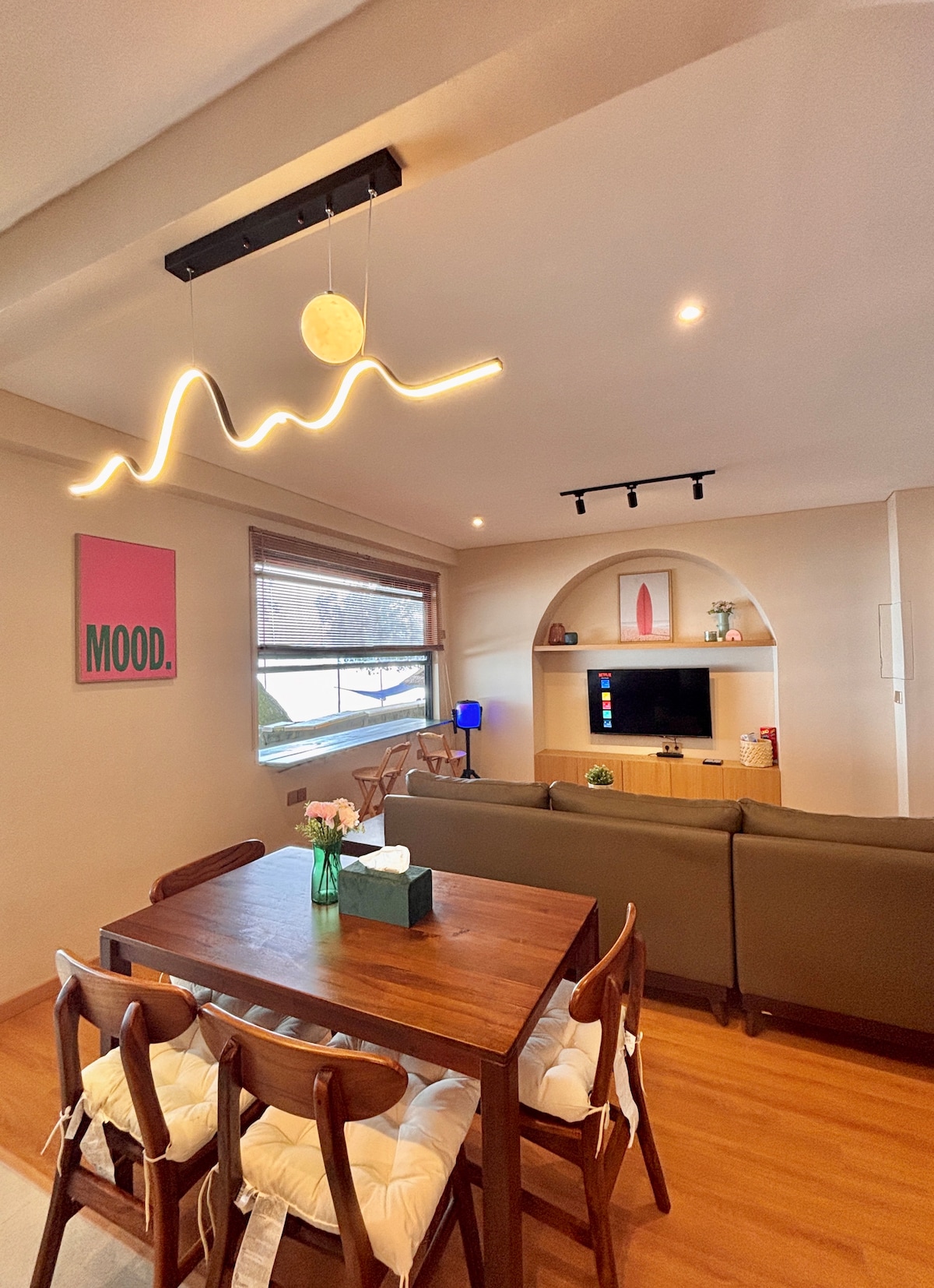 A modern living space is shown, featuring a wooden dining table surrounded by chairs. A comfortable sofa is positioned nearby, facing a wall-mounted television. Soft lighting accents the room, and a playful artwork is displayed on the wall. Large windows provide natural light.