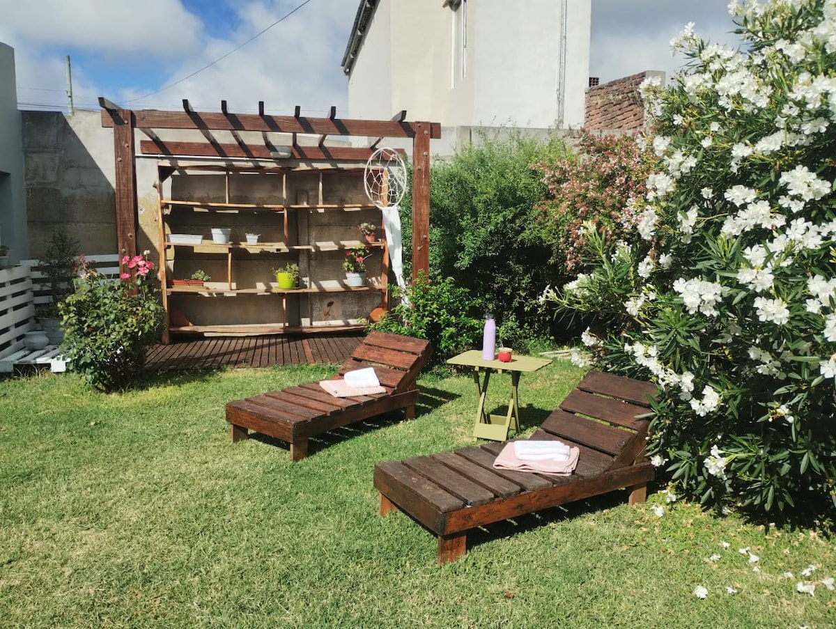 A peaceful garden area is presented, featuring two wooden lounge chairs positioned on lush green grass. A small table holds a candle, with towels neatly placed on the chairs. Flowering bushes surround a wooden shelf adorned with plants, adding a touch of greenery.
