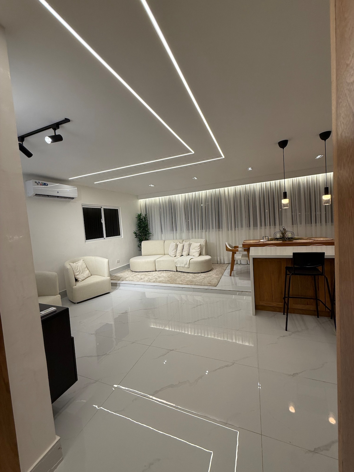A modern living area features a light-colored sofa and armchair arranged around a soft rug. A sleek kitchen bar with high stools is visible in the background. Bright LED lighting highlights the spaciousness, complemented by the gentle glow of natural light from the windows.