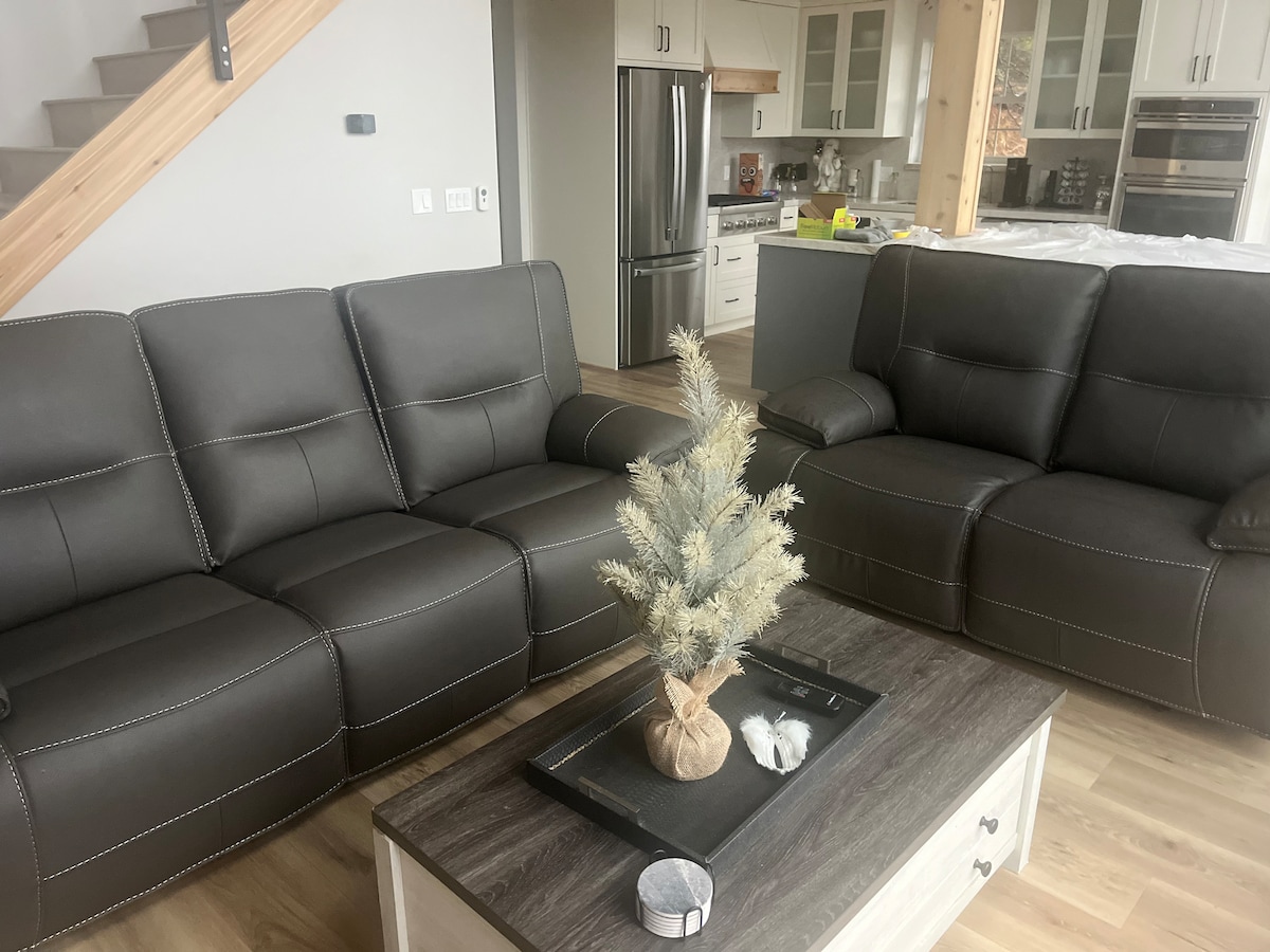 A comfortable living area features two dark leather sofas arranged around a wooden coffee table. A small decorative tree is placed in a woven vase atop the table, with a candle and a few decorative items present. The background reveals a modern kitchen with stainless steel appliances.