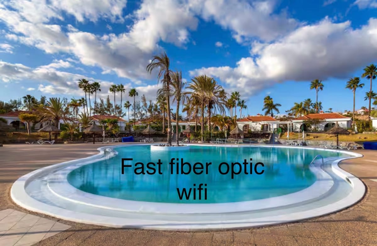 A spacious swimming pool is surrounded by palm trees, with lounge chairs arranged alongside. The clear water reflects the bright blue sky dotted with clouds. The tranquil atmosphere is enhanced by the inviting layout of the pool area.