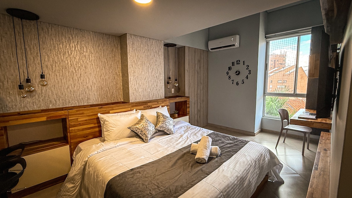 A spacious bedroom features a king-size bed with premium linens and decorative pillows. A wooden headboard adds warmth, while a modern clock and light fixtures enhance the contemporary design. A desk and chair are positioned by the large window, offering views of the cityscape.