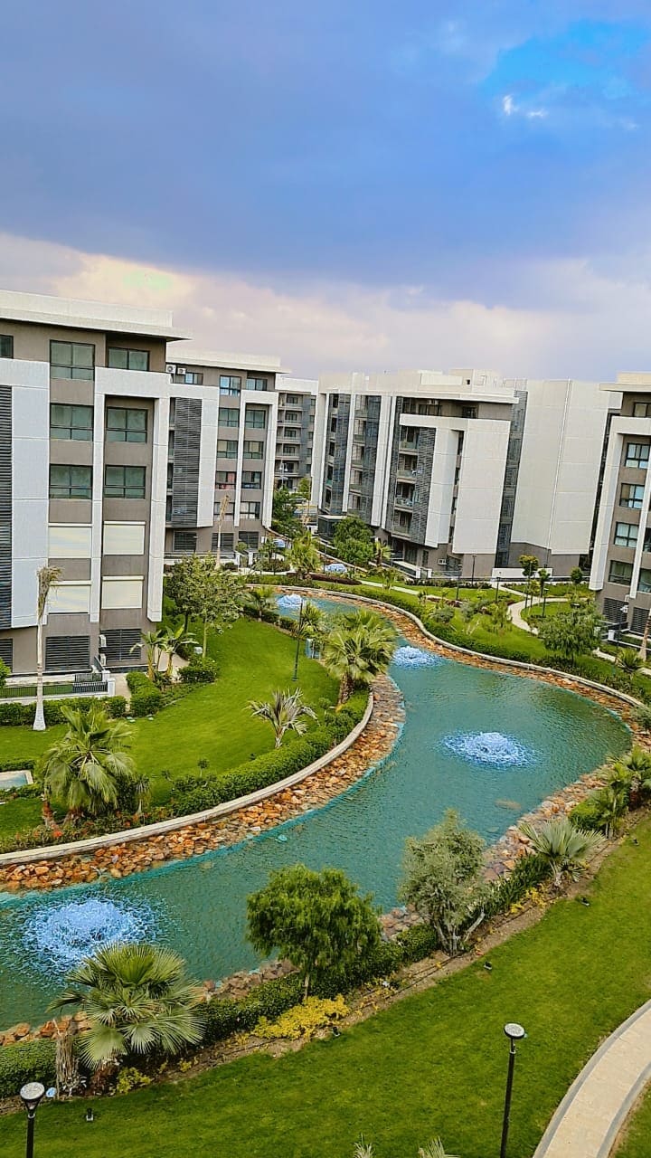 The image captures a serene view of landscaped green spaces featuring a winding waterway. Modern apartment buildings encircle the lush area, with palm trees and manicured shrubs enhancing the natural beauty. Soft blue water features are visible in the stream, inviting a sense of tranquility.