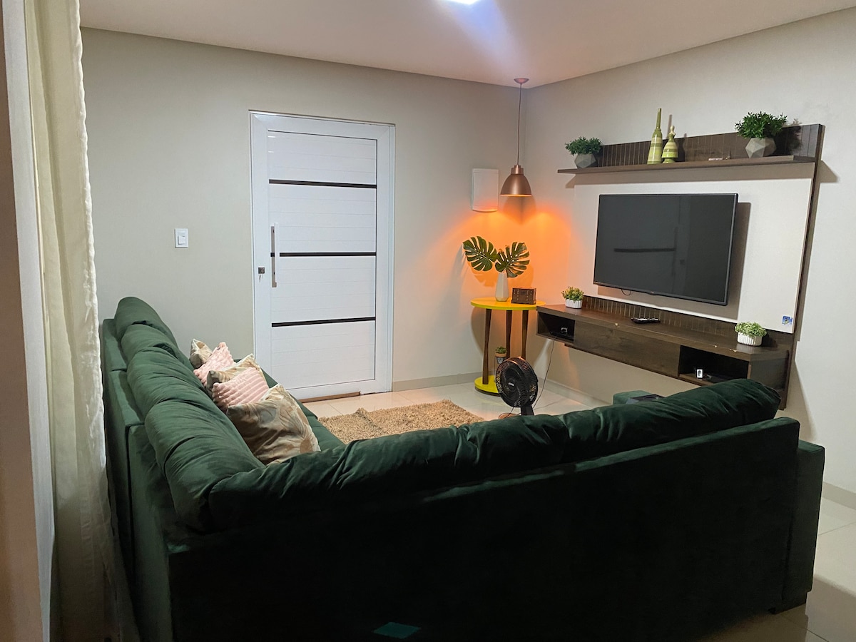 A spacious living area features a plush green sofa adorned with decorative pillows. A television is mounted on a wooden shelf, accompanied by potted plants. Natural light enters through a window, complementing the warm tones from a pendant light.