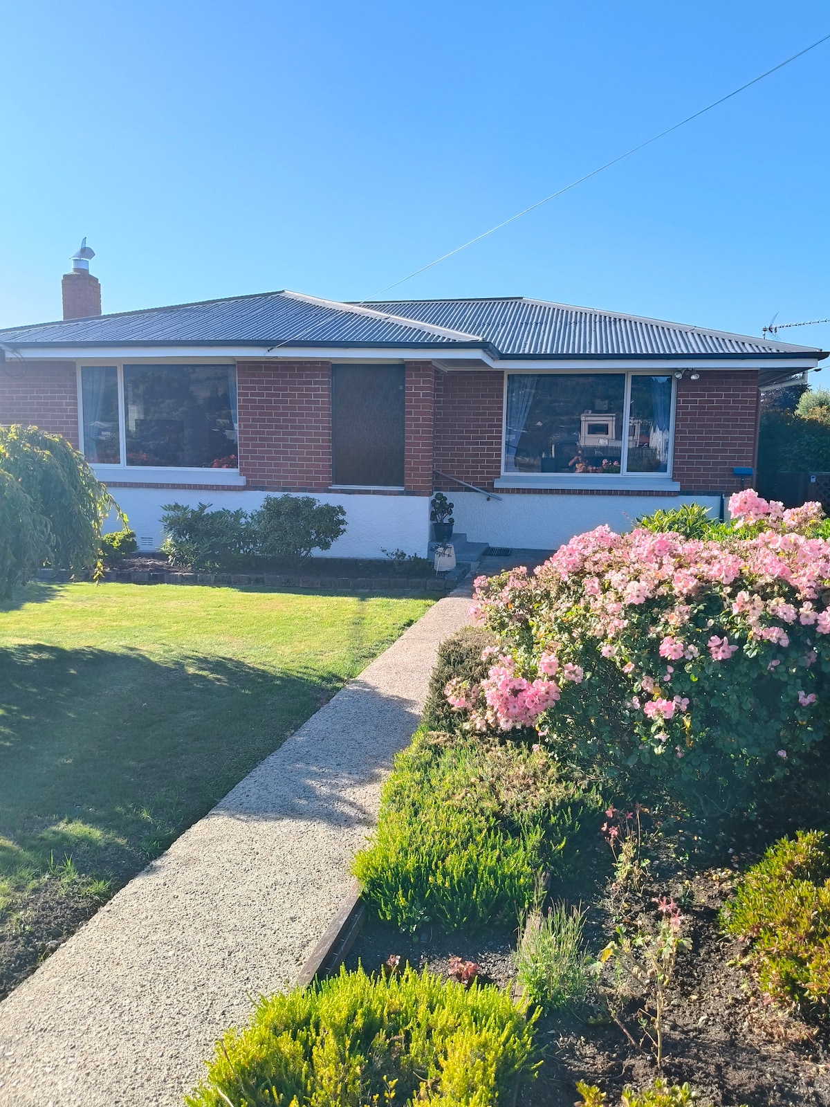 A charming house is visible, featuring a red brick exterior and a welcoming entrance. Lush greenery surrounds the property, with flowering shrubs in soft pink hues in the foreground. The clear blue sky provides a bright backdrop to this inviting residential setting.