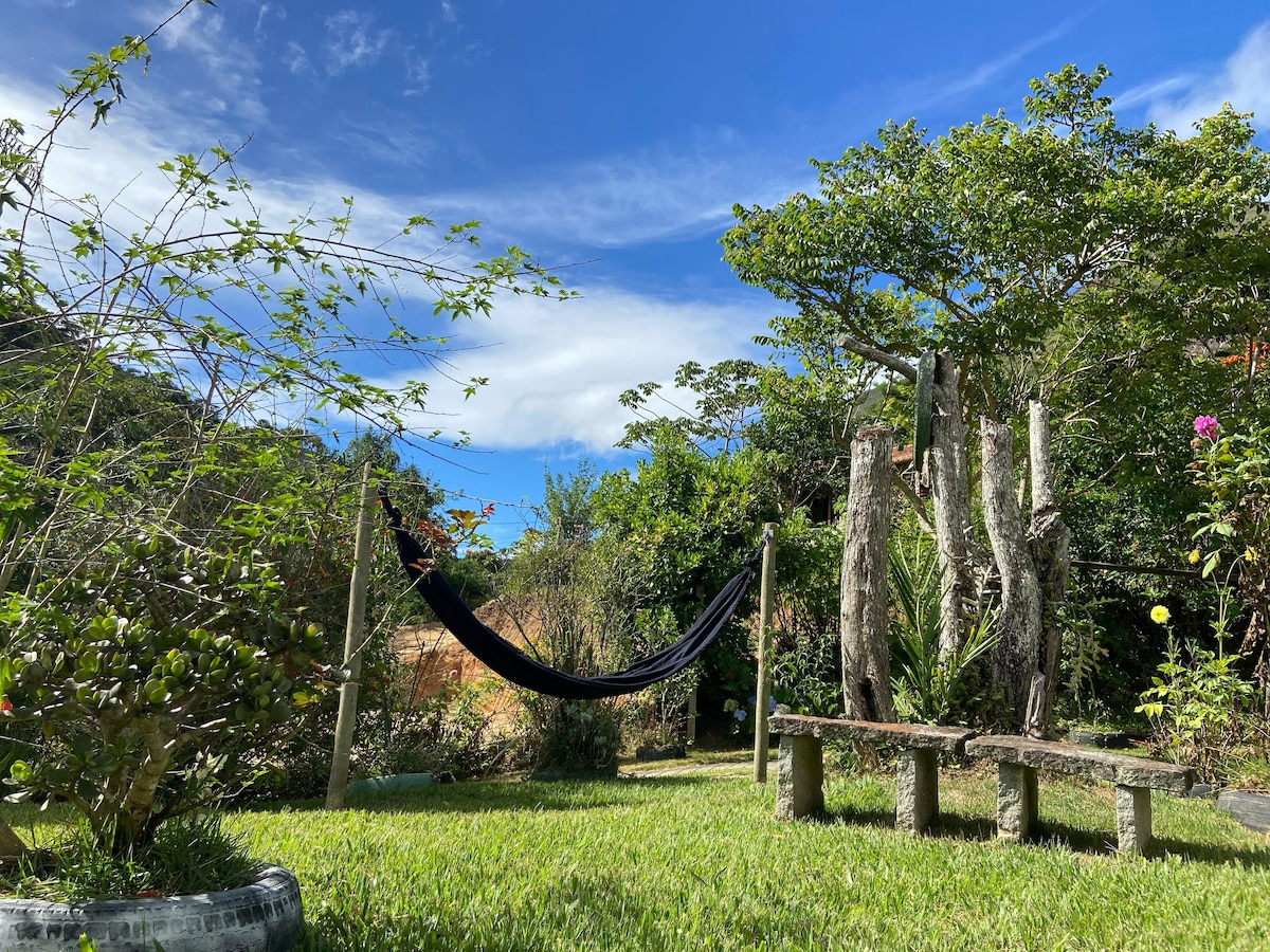 A tranquil outdoor space is presented, featuring a black hammock strung between wooden posts. Natural greenery surrounds the area, with vibrant flowers and trees visible in the background. A rustic wooden bench sits on a grass-covered surface, inviting relaxation and connection with nature.