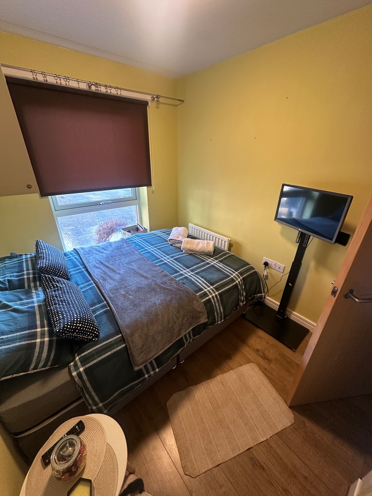 A small cozy bedroom features a spacious bed with a plaid comforter and neatly arranged pillows. A mounted television stands on a sleek black stand, while a soft area rug lies beneath a bedside table. Natural light filters through a window with dark blinds.