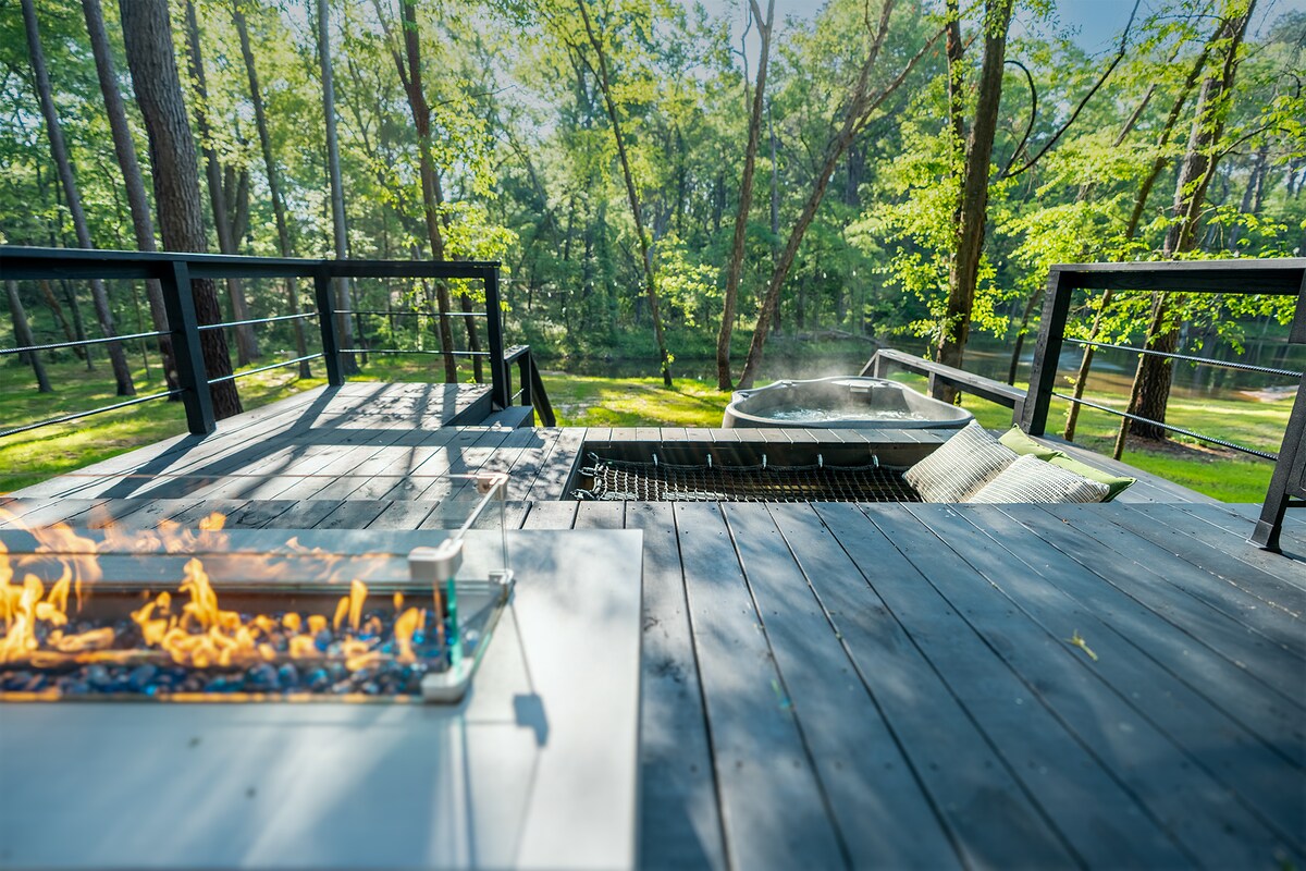 A spacious deck overlooks a lush, green landscape, featuring a private hot tub gently steaming in the distance. A fire pit with a decorative flame is positioned nearby, creating a cozy ambiance for outdoor gatherings. Natural light filters through the surrounding trees.